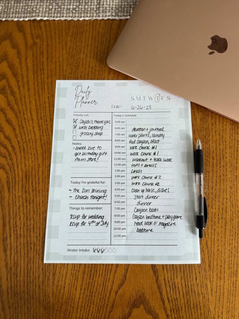 Flatlay image of a printed time blocking planner with handwritten notes, a pen, and a rose gold MacBook on a wooden kitchen table—showing a practical way moms can have a productive day. 
